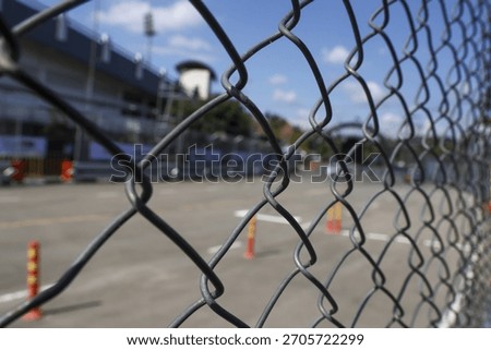 Restricted background view of an empty racetrack and grandstand seen through metal chain link fence under blue sky, creating feeling of anticipation and confinement