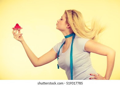 Restrictive Diet, Temptation, Losing Weight Concept. Woman Holding Strawberry Sweet Cupcake Having Measure Tape Around Her Neck. Studio Shot On Yellow Background