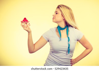 Restrictive Diet, Temptation, Losing Weight Concept. Woman Holding Strawberry Sweet Cupcake Having Measure Tape Around Her Neck. Studio Shot On Yellow Background