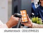 Retail employee scanning smartphone with Black Friday discount coupon while serving customer at cash register desk, close up. Clothing store marketing promotion during seasonal sales