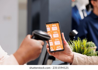Retail employee scanning smartphone with Black Friday discount coupon while serving customer at cash register desk, close up. Clothing store marketing promotion during seasonal sales - Shutterstock ID 2311594121