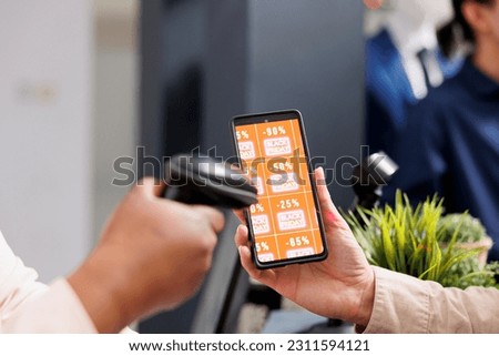 Retail employee scanning smartphone with Black Friday discount coupon while serving customer at cash register desk, close up. Clothing store marketing promotion during seasonal sales