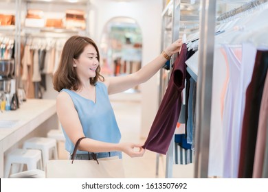 Retail Shopping Concept, Beautiful Asian Thai Girl Chooses Clothes In A Shopping Mall, Brown Hair Smiling Women Trying On A Blouse, She Is Very Happy After Work In The Business Park Near Her Office   