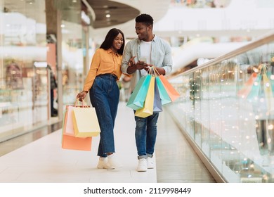 Retail And Technology. Happy African American couple using smartphone, cheerful guy holding cell and shopper bags, lady pointing finger at screen. Young family walking in city mall, full body length