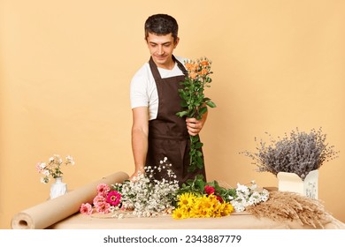 Retailer of fresh flora. Beautiful flower compositions. Creative garden arrangements. Young male in brown apron working in flower shop standing isolated over beige background - Powered by Shutterstock