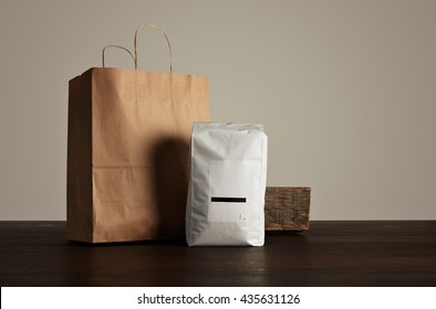 Retailer Merchandise Pack: Big Hermetic Pouch White With Blank Label Presented Near Of Craft Paper Bag And Rustic Wooden Brick On Red Table, Isolated On Gray. Coffee Or Tea Distribution