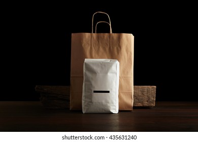 Retailer Merchandise Pack: Big Hermetic Pouch White With Blank Label Presented In Front Of Craft Paper Bag And Rustic Wooden Brick On Red Table, Isolated On Black. Coffee Or Tea Distribution