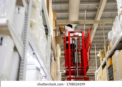 Retailer Warehouse Stock Of Produce Food And Products, Worker Employee Man Upon Human Lifter Machine Checking Item And Products Restocking And Replacing, With Big Cardboard Box Stacking A High Shelf