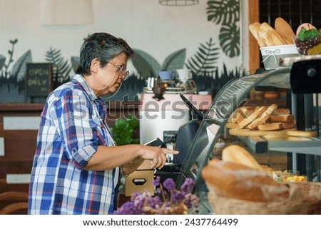 Retired Asian female barista wearing a plaid shirt Check the amount of bread and bakery items from the tablet. will be enough for the next day. small coffee shop and family businesses of retired women
