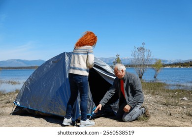 Retired couple enjoying a sunny day by a lake, setting up their tent during a delightful camping trip filled with adventure and relaxation - Powered by Shutterstock