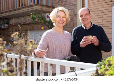 Retired Couple Outside Their House On A Terrace With Gardening Instruments And A Cup Of Hot Beverage