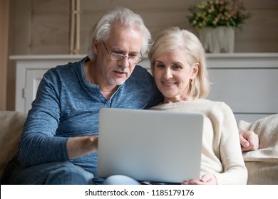 Retired Couple Using Laptop Together Sitting On Sofa, Smiling Senior Middle Aged Man And Woman Read Internet News Talking Shopping Online At Home, Elderly Family Customers With Computer Lifestyle