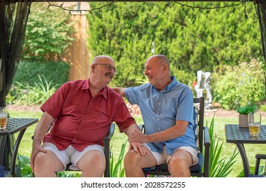 A Retired Gay Married Couple In Love, Relax In Their Back Yard And Laugh As They Sit And Talk.  One Has His Hand On His Husband's Knee While The Other Has His Hand On His Husband's Shoulder.