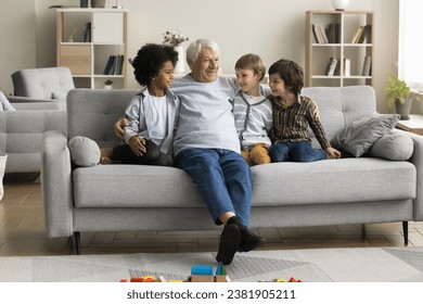 Retired great-grandfather talks to three multiethnic little great-grandchildren sitting together on couch at home enjoy conversation, older relative tell stories and memories, teach, share experience - Powered by Shutterstock