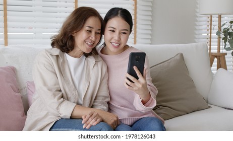 Retired Healthy Mom And Girl Two People Hold Phone Waving Hand Greeting Selfie Look At Camera On Webcam App At Relax Comfort Sofa Couch Living Room In Health Care Quarantine Social Distance Isolate.