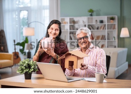 Retired Indian asian couple do paperwork at home with laptop, piggy bank, currency notes, documents