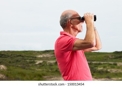 Retired man with beard and glasses using binoculars outdoors in grass dune landscape. Wearing pink t-shirt. - Powered by Shutterstock