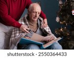 Retired man looking photo album with old photos sitting on armchair while his son taking care of pensioner covering dad with blanket. Copy space