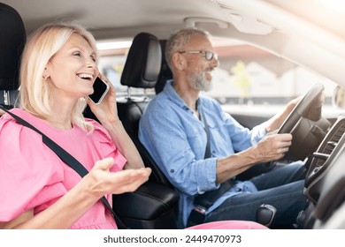 A retired married couple in their car, with the woman having a cheerful phone conversation as the senior man drives with a happy demeanor - Powered by Shutterstock