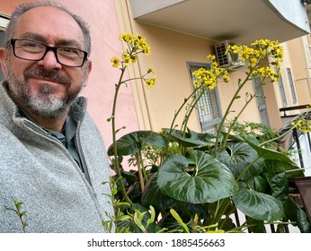 Retired Middle Aged Italian Man With Short Hair And Beard And Glasses Poses In A Selfie Next To A Plant With Large Leaves On A Balcony