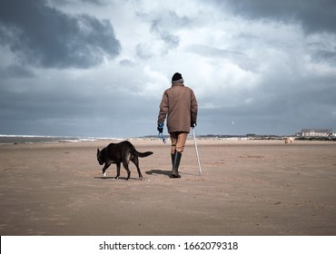 A Retired Old Man With A Walking Stick, Walking His Dog On A Beach. Dogs Are Mens Best Friend And  Loyal Companions For Older People Who Are Alone. Humans And Animals Together.