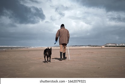 A Retired Old Man With A Walking Stick, Walking His Dog On A Beach. Dogs Are Mens Best Friend And  Loyal Companions For Older People Who Are Alone. Humans And Animals Together.