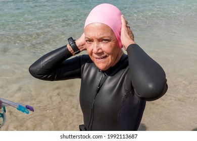 Retired Older Woman Dressed In A Wetsuit Putting On A Pink Swim Cap Looking At The Camera.