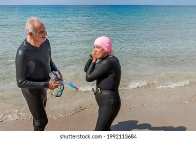 Retired Older Woman In A Wetsuit Putting On Her Swimming Cap While Her Husband Looks On.