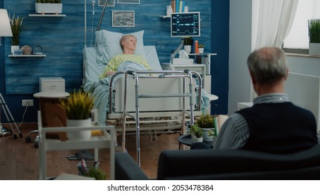 Retired Patient With Disease Sitting In Hospital Bed With Heart Rate Monitor And IV Drip Bag In Nursing Home. Ill Woman Waiting For Healing Treatment While Senior Man Assisting Sick Wife