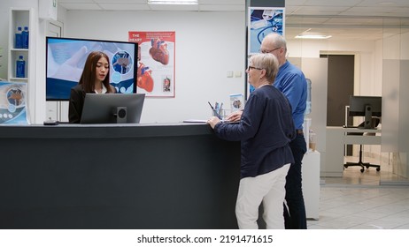 Retired people doing medical report with receptionist in hospital waiting room, woman at reception desk helping patients with checkup appointment. Assistant working in healthcare center. - Powered by Shutterstock