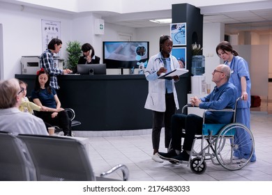 Retired Person Using Wheelchair Being Helped By Professional Nurse Talking With African American Doctor For Appointment. Older Man Living With Disability In Hospital Reception With Diverse People.