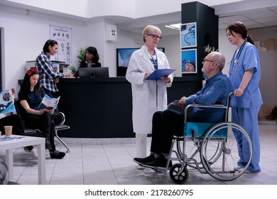 Retired Person Using Wheelchair Talking With Senior Doctor For Appointment While Being Helped By Professional Nurse In Private Clinic. Older Man Living With Disability In Busy Hospital Reception.