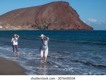 Retired Senior Couple Walking On The Shore With Their Feet In The Water While Holding Their Hats Due To The Wind. Happy Active People On Winter Holidays By The Sea