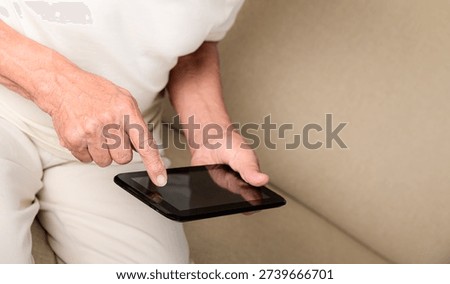 Retired senior man using touchscreen tablet sitting comfortably on couch at home.
