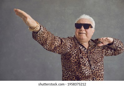 Retired senior man vibing to hype pop music. Portrait of funny rich white haired old grandpa in cool glasses and leopard patterned party shirt flexing, dancing and having fun on gray studio background