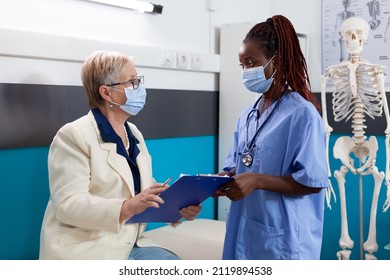 Retired Senior Woman Patient With Protective Face Mask Against Coronavirus Holding Clipboard With Medical Documents Signing Medication Prescription Paper In Hospital Office. Medicine Concept