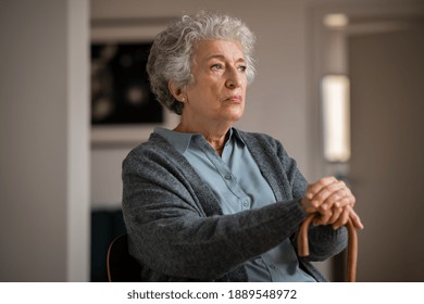 Retired unhappy woman at home. Lonely serious senior woman holding wooden walking stick and looking through the window. Moody and upset grandmother sitting on couch in nursing home. - Powered by Shutterstock