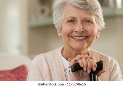 Retired woman with her wooden walking stick at home. Happy senior woman relaxing at home holding cane and looking at camera. Smiling grandmother sitting on couch.