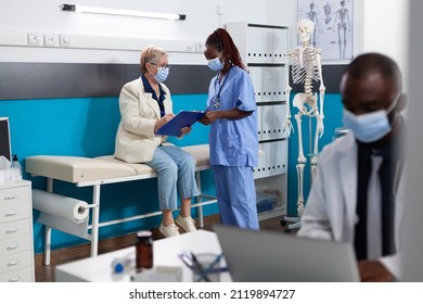 Retired Woman Patient With Protection Face Mask Signing Medical Documents During Medical Appointment In Hospital Office. Therapist Nurse Explaining Sickness Diagnosis Discussing Medication Treatment