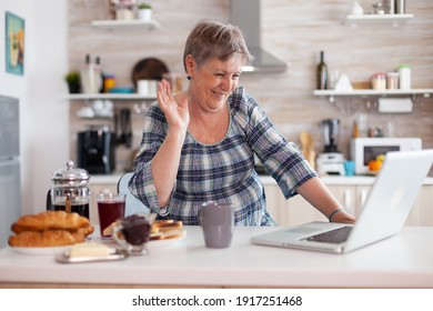 Retired Woman Waving During Online Meeting With Family On Video Call Using Laptop In Kitchen Having Breakfast. Elderly Person Using Internet Online Chat Tech, Pc Webcam For Virtual Conference Call