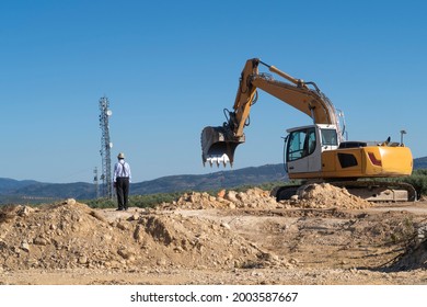 A Retiree Looking At A Construction. A Work On A Ground Of Disturbed Earth: An Excavator With Chains Preparing The Bucket To Catch Earth. In The Background A Cell Tower And A Mountain With Vegetation.