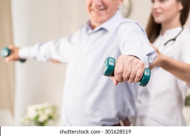 Retiree Training With Dumbbells Assisted By Young Physiotherapist