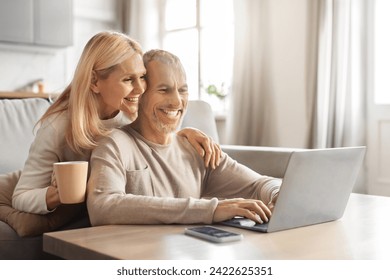 Retirement and internet fun concept. Excited smiling senior spouses using modern laptop computer, websurfing or watching film online, resting on sofa at home, embracing, drinking coffee, free space - Powered by Shutterstock