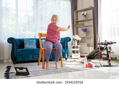 Retirement woman sitting on a wooden chair, Pushing hand to the opposite side, watch at tablet during workout with training online at home, senior exercise concept