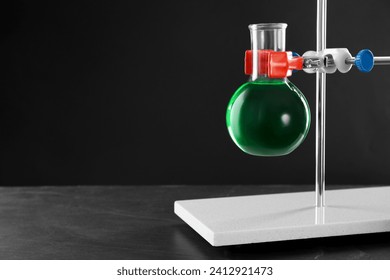 Retort stand and laboratory flask with liquid on table against black background, closeup. Space for text