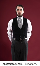 Retro 1900 victorian fashion man with beard wearing black gilet and grey tie. Studio shot against red wall.