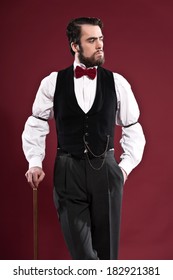 Retro 1900 victorian fashion man with beard wearing black gilet and red bow tie. Holding a walking stick. Studio shot against red wall.