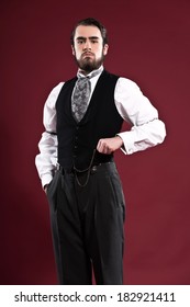 Retro 1900 victorian fashion man with beard wearing black gilet and grey tie. Holding pocket watch. Studio shot against red wall.