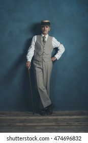 Retro 1920s dandy in suit standing with cane in front of gray wall.