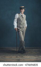 Retro 1920s dandy in suit standing with cane in front of gray wall.